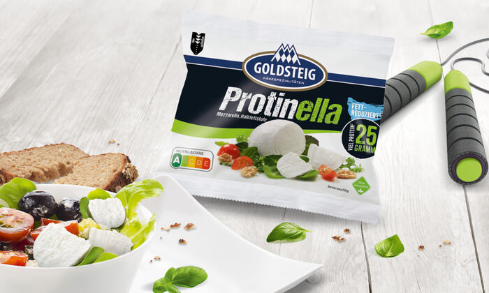 Startseite (GOLDSTEIG): Proteinkäse Protinella von GOLDSTEIG. Verpackung drapiert auf grauem Holztisch mit einem leckeren Salat und einem Springseil.